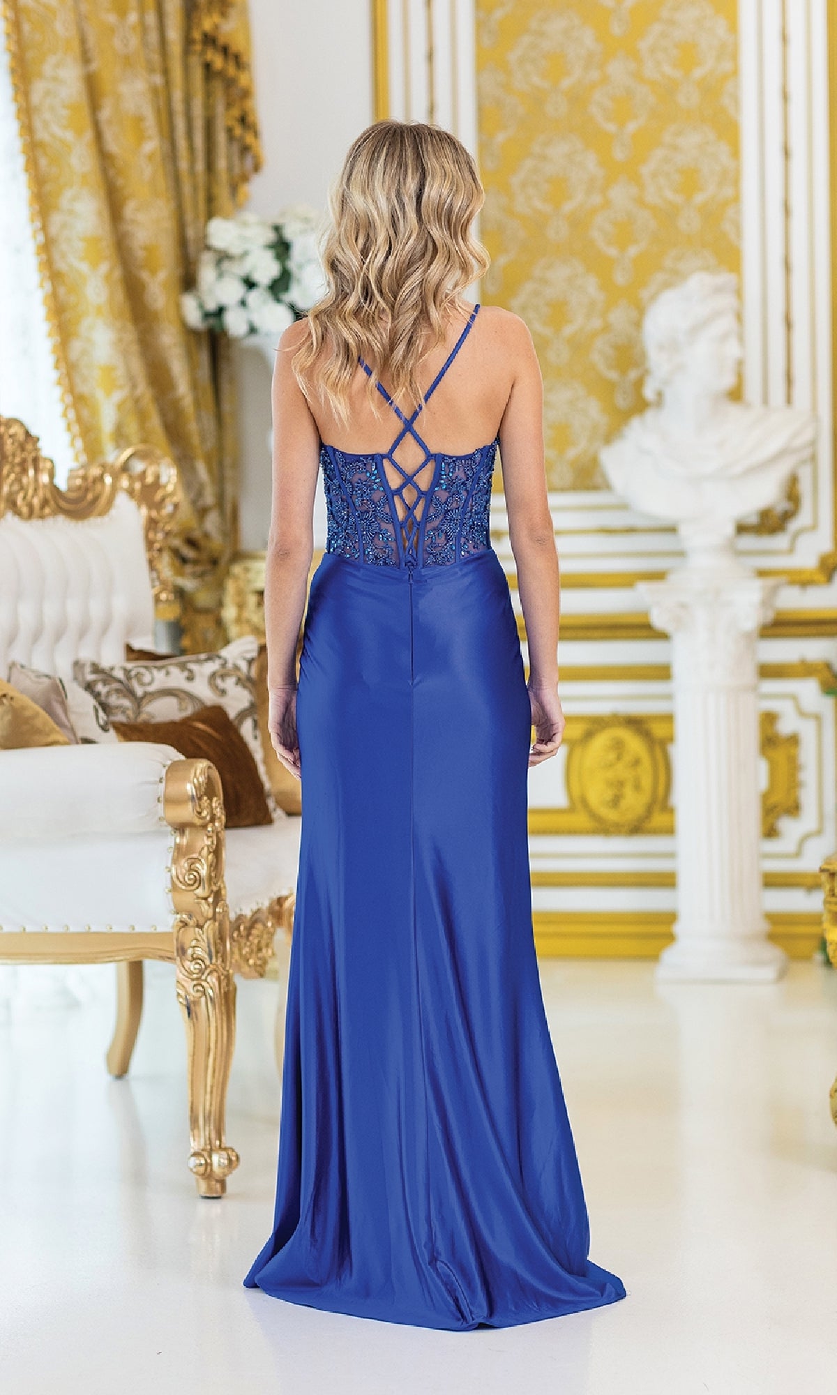 A woman with wavy blonde hair wears the Drape-Front Long Prom Dress: Dancing Queen 4439, which features a beaded bodice and crisscross straps, as she stands in an ornate room with gold decor and classic furnishings.